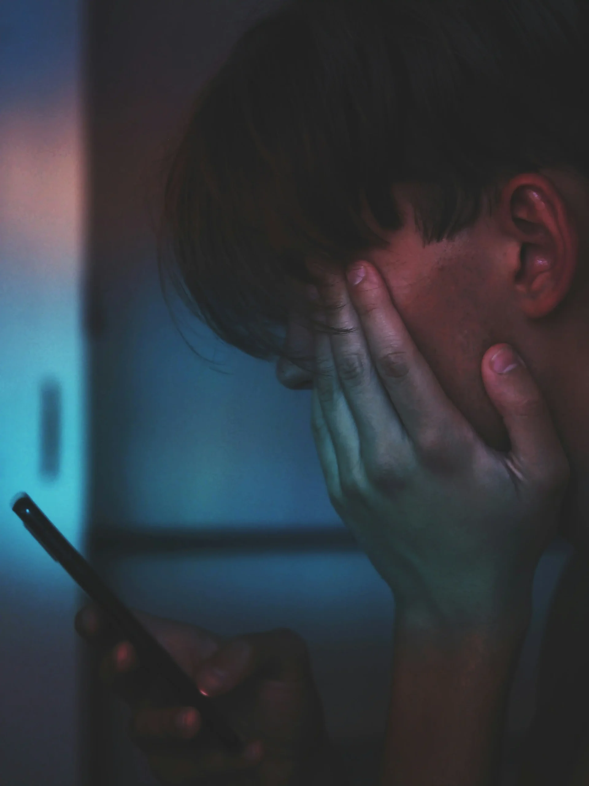 A person holding their face while looking at a smartphone glowing in a dark room