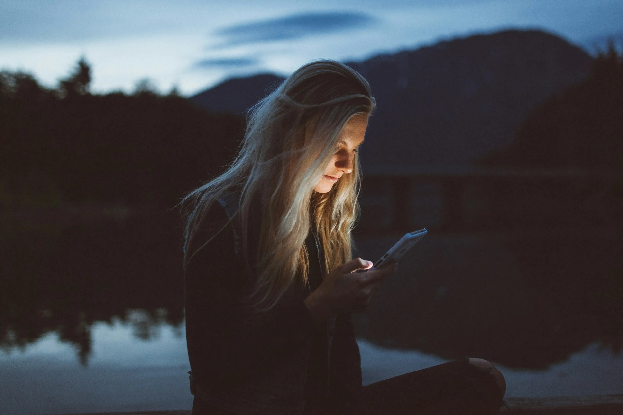 A woman sitting by a lake at dusk, looking at her phone