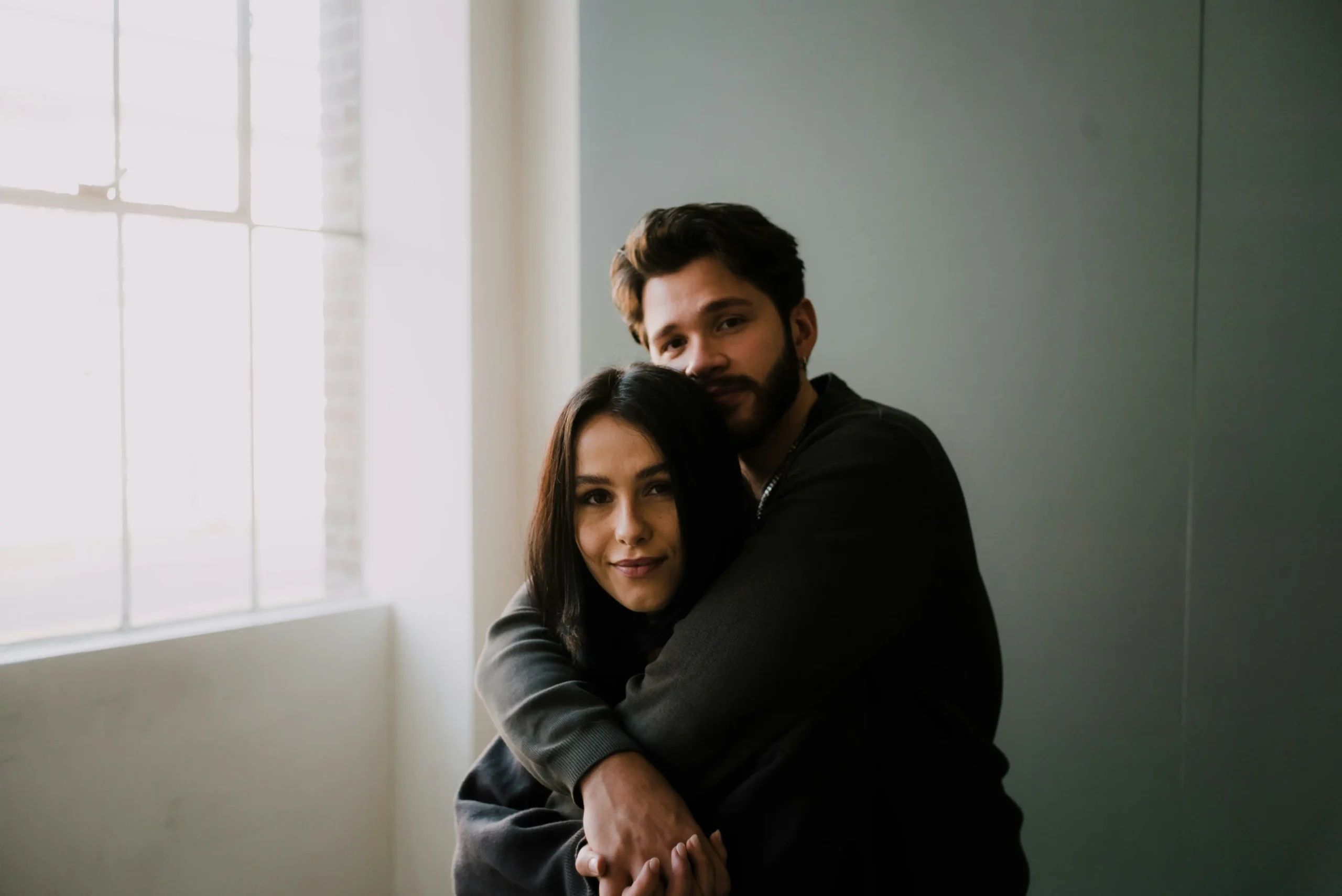 A couple embracing warmly by a window, the man wrapping his arms around the woman