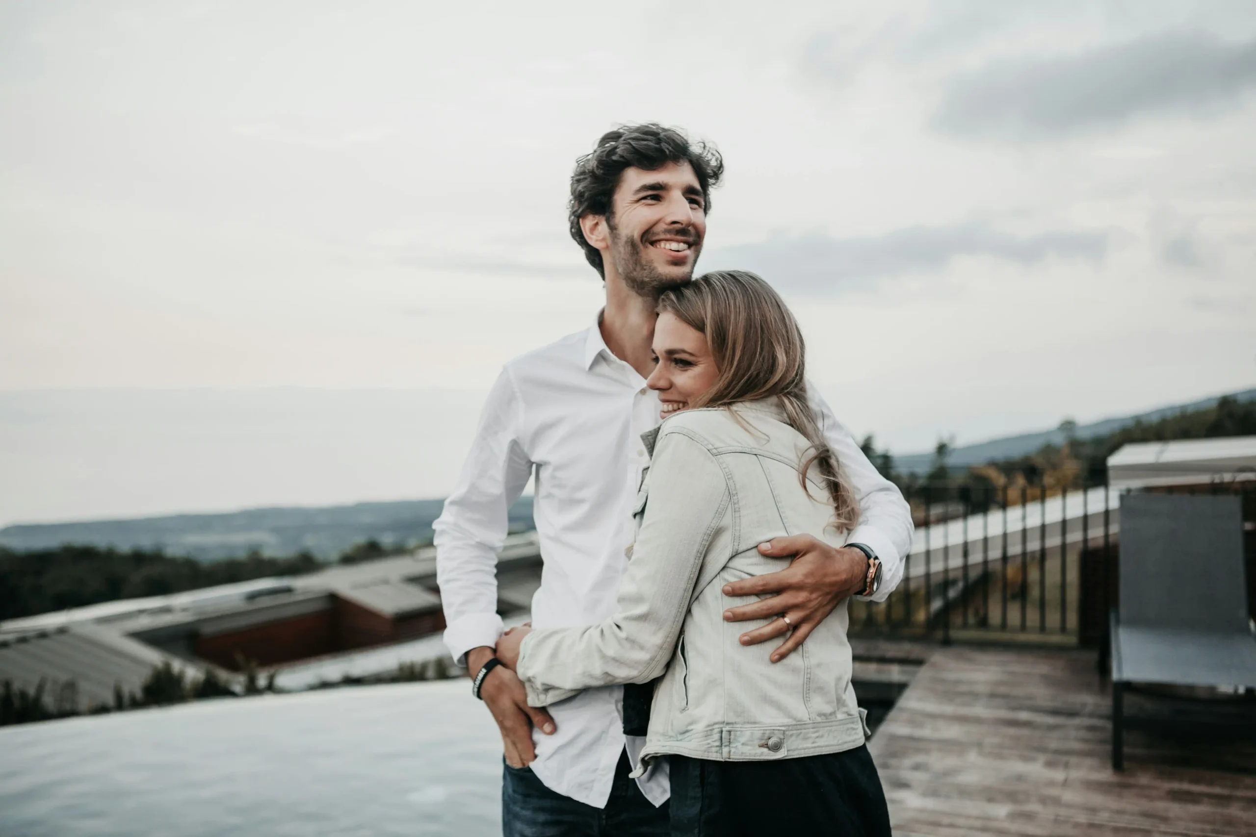 A couple embracing outdoors with a scenic overlook behind them, both smiling