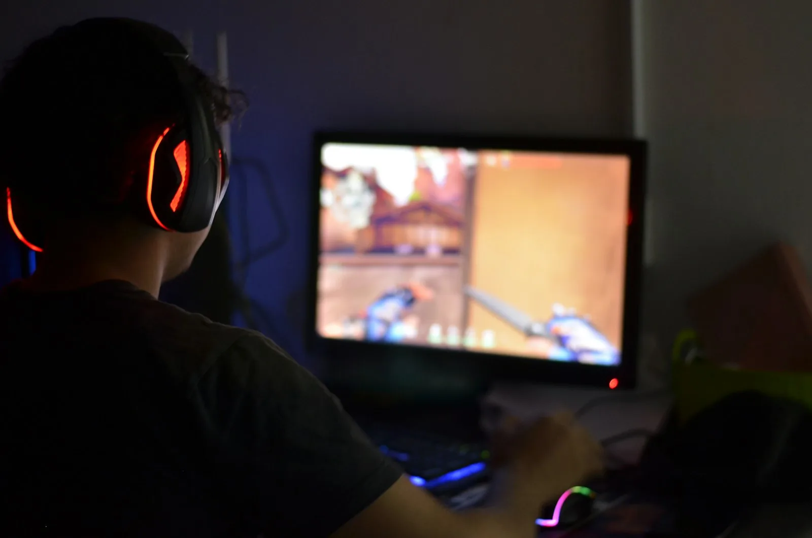 A young man wearing gaming headphones, seen from behind, playing a video game lit only by his monitor in a dark room