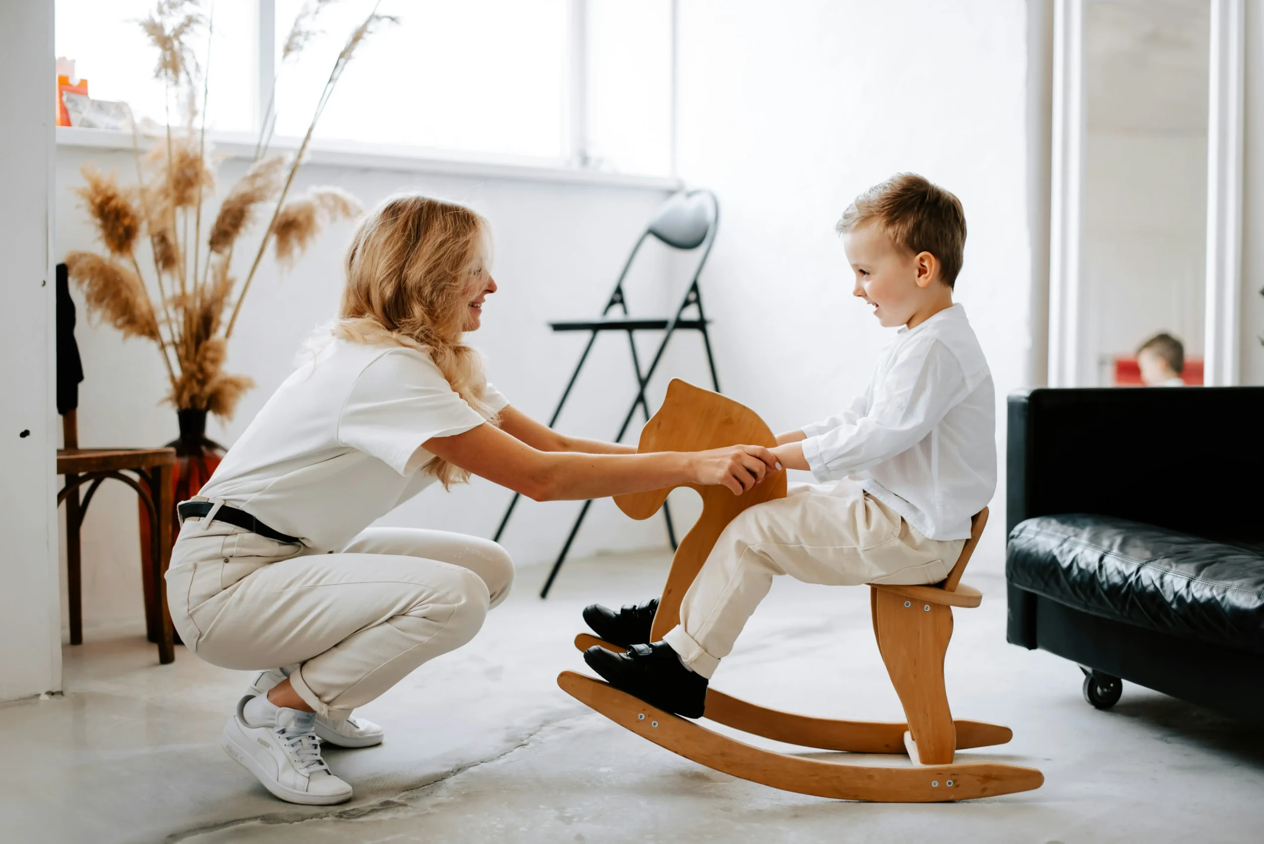 A mother crouching down to play with her young son on a wooden rocking horse, both smiling