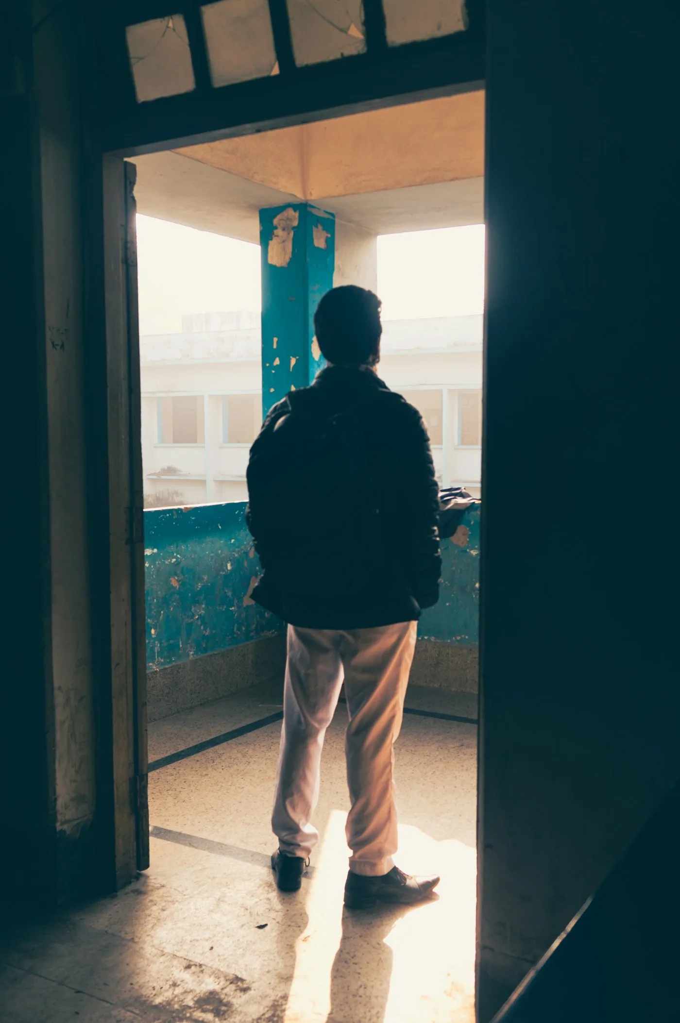 A young man standing at a doorway threshold, backpack on, looking out into the light beyond