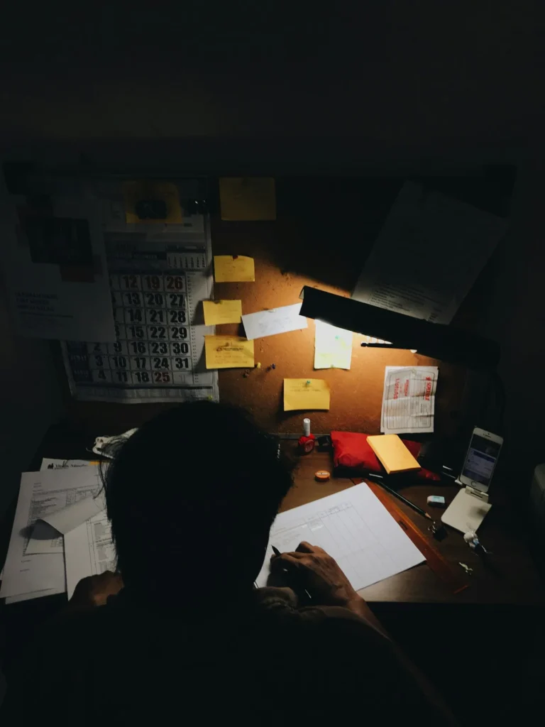 A person at a cluttered desk lit by a lamp in a dark room
