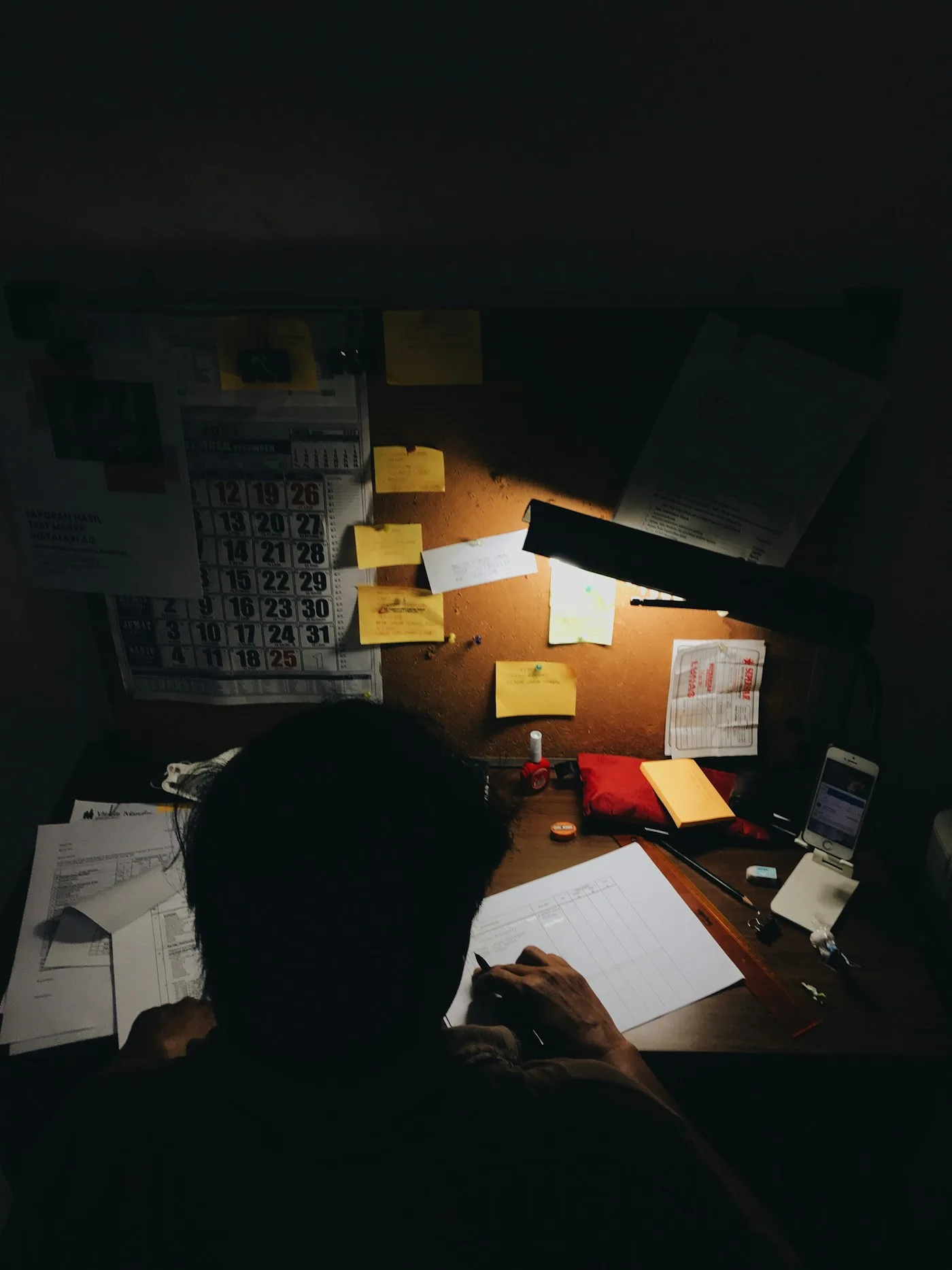 A person at a cluttered desk lit by a lamp in a dark room