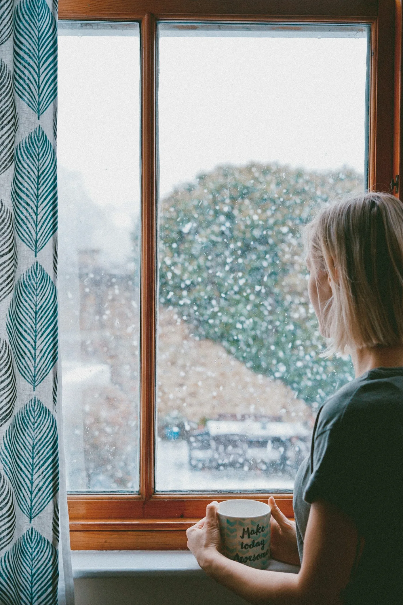 A woman holding a mug and looking out a rainy window