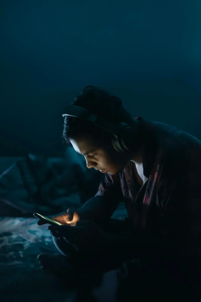 A young man sitting alone in a dark room at night, illuminated only by his phone screen