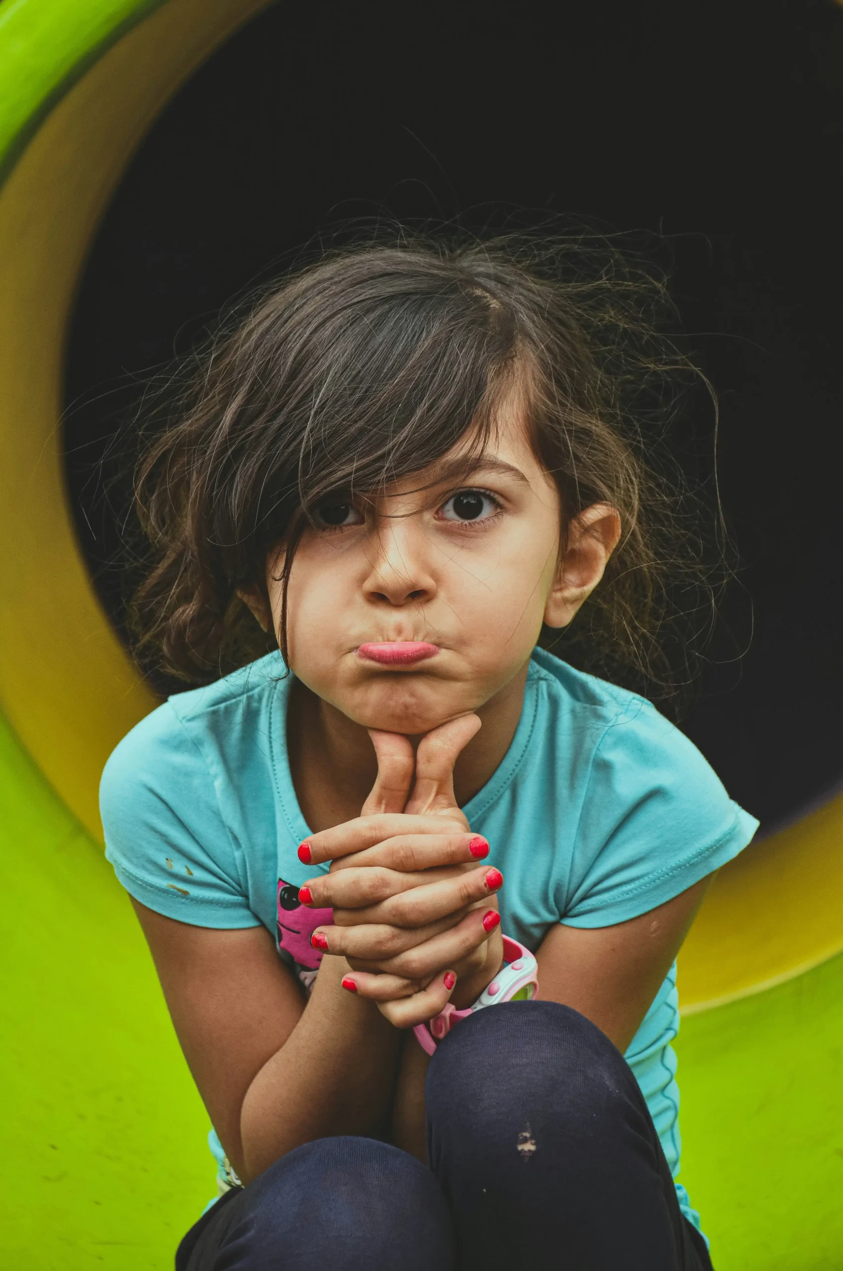 A young girl sitting on playground equipment with a sad, withdrawn expression, chin resting on her clasped hands