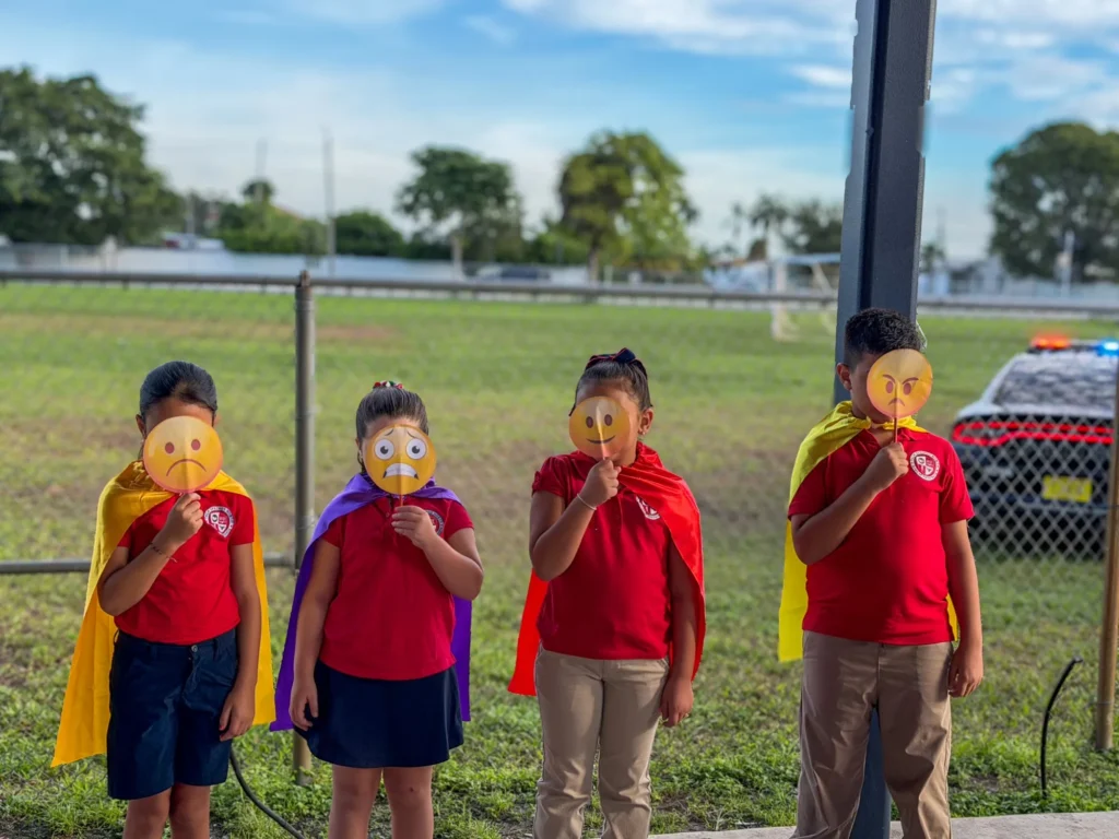 Students in capes hold emoji faces showing different feelings at the Behavioral Aid Solutions mental health fair station at Palm Springs North K-8 Center